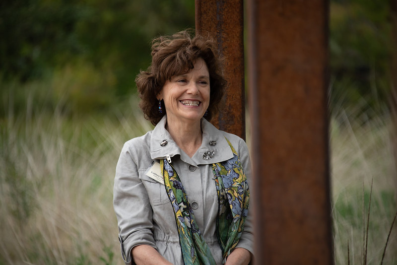 Image of Kathleen Socolofsky smiling, wearing a gray cinch-waisted blazer and scarf while leaning against a corten steel column.