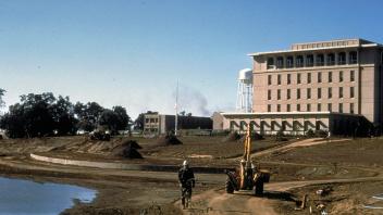 Cyclist on dirt path past excavator beside drained lake and large beige building