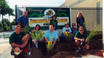 Eight adults wearing leis smile by a colorful welcome sign and potted plants outdoors