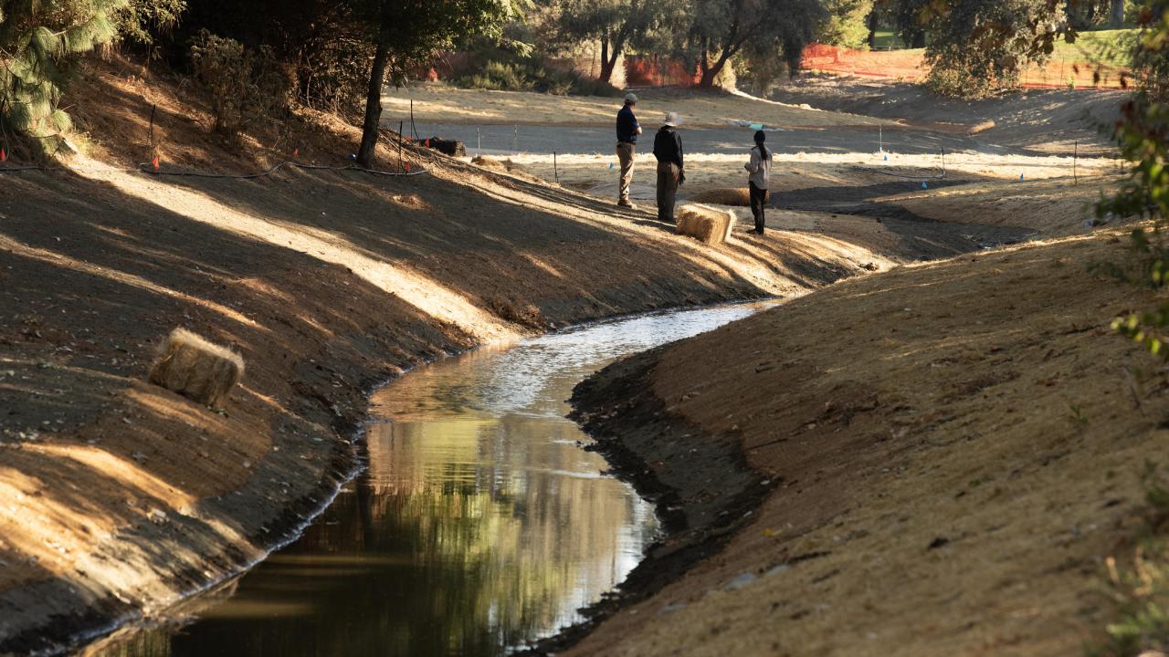 In the foreground, a narrow, curving portion of the Arboretum Waterway is shown with water while three people look in the opposite direction to a larger area of the Waterway.