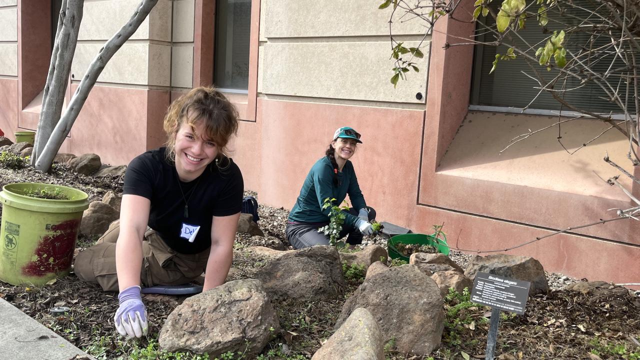 Two volunteers kneeling by a building, weeding a rock garden, one smiling at camera