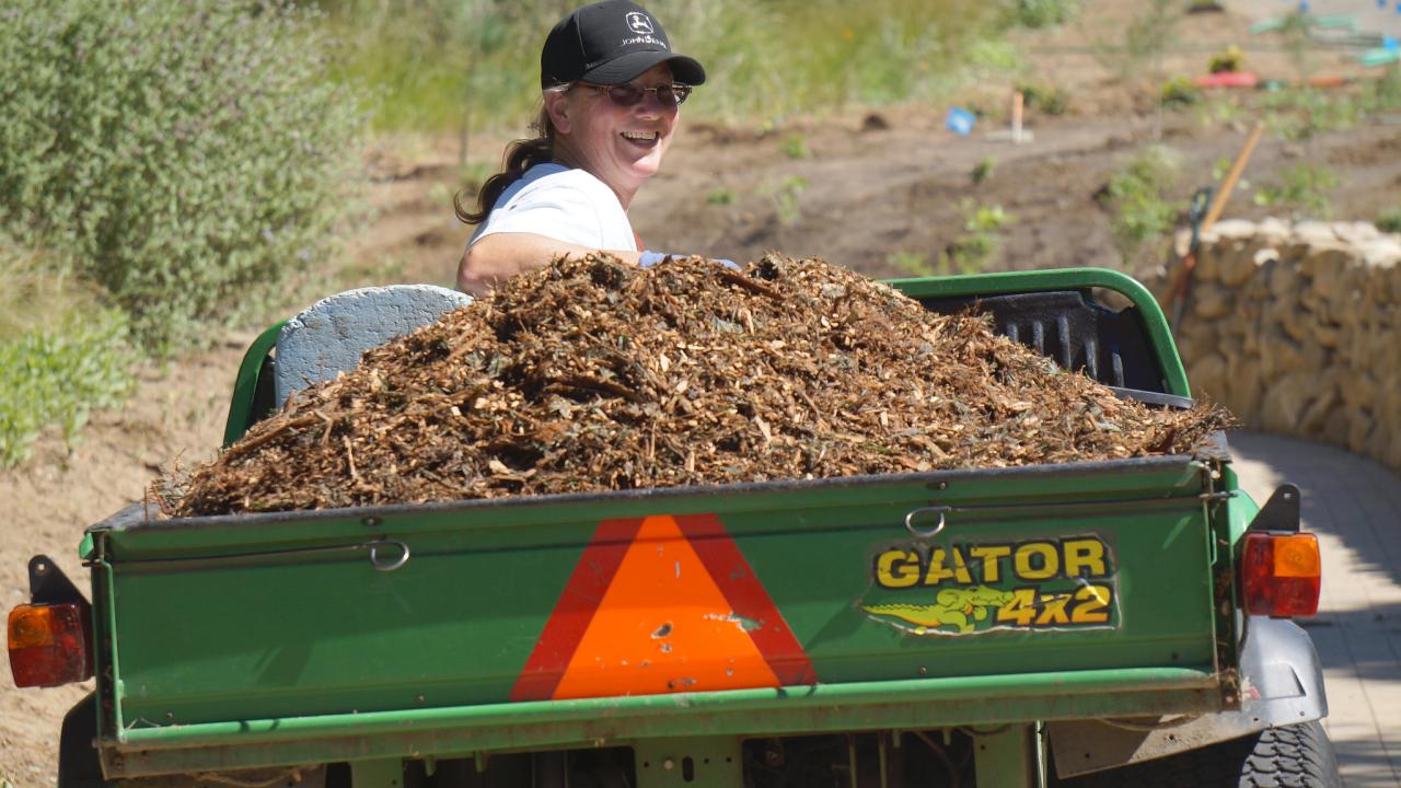 Image of woman in a baseball hat looking over her shoulder while reversing a small cart with a pick-up bed filled with mulch.