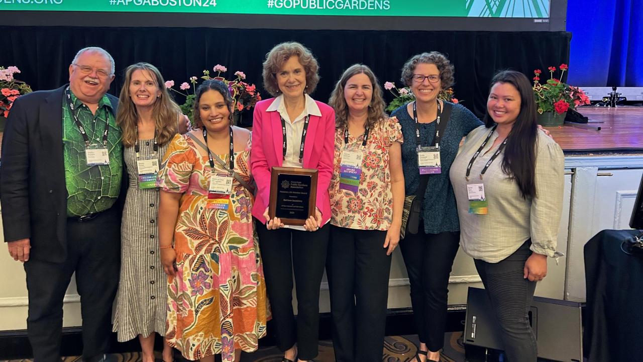 Kathleen Socolofsky holds an award, standing in the center of a group of seven people in front of a stage.