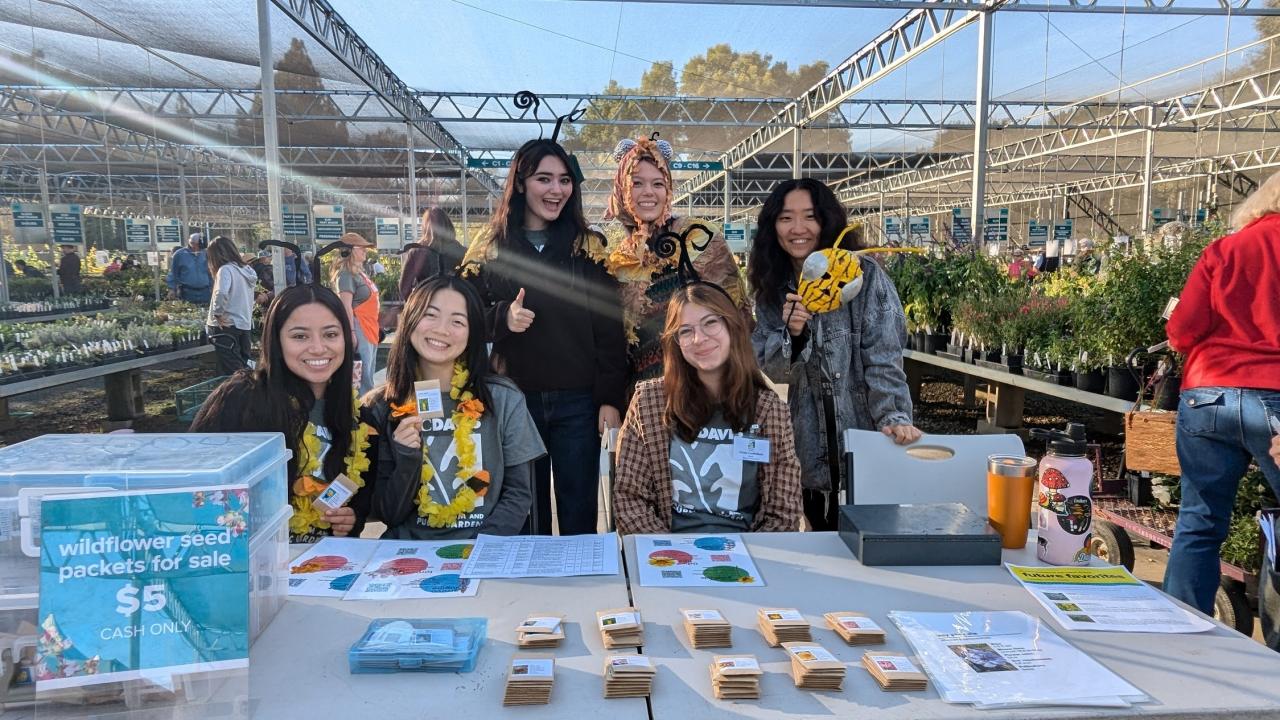 Group of student interns pose, smiling behind a vendor table in the Arboretum Teaching Nursery