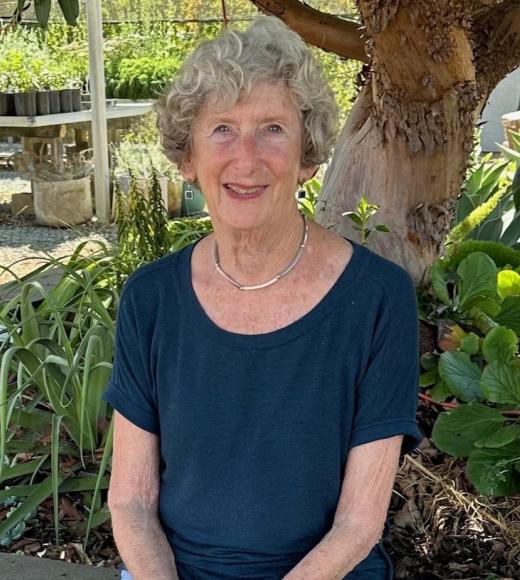 woman sitting down wearing navy blue blouse. green foliage and tree behind her