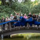 Image of a line of people on an arched bridge celebrating by cutting a large blue ribbon on the side of the bridge railing.
