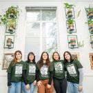 Five students wearing Arboretum and Public Garden t-shirts smile in front of a wall decorated with hanging plants.