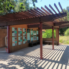 A wide-angle outdoor view of the Waterway Education Pavilion near Putah Creek Lodge at UC Davis. The small open-air structure has a wooden pergola-style roof casting striped shadows on the ground. A wall of educational signs is visible inside, along with built-in wooden benches. The structure is surrounded by native vegetation, with a wood fence and nature trail nearby.